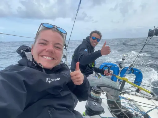 Hugo Dhallenne et sa coéquipière souriants sur un voilier, pouces levés, en pleine mer sous un ciel couvert.