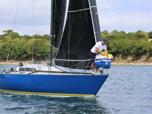 Un homme prépare le bateau, une voile noire déployée, sur fond de côte boisée. Il semble être le **half toner** de l'équipage.
