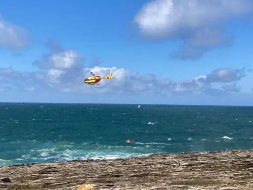 Un hélicoptère jaune et rouge survole une mer agitée, cherchant une personne disparue en mer. Un petit bateau rouge est visible au loin.