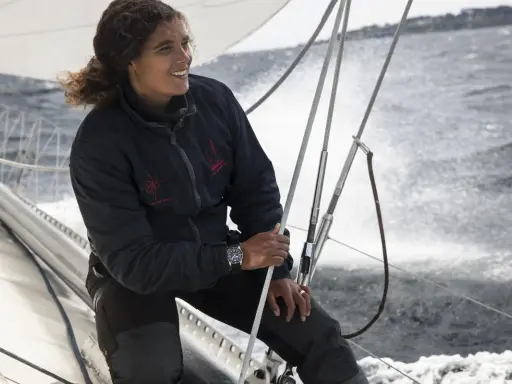 Marie Tabarly, souriante, tient la barre d'un voilier, le vent dans ses cheveux, naviguant sur une mer agitée.