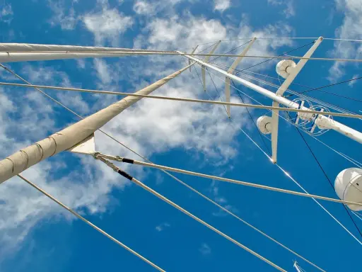 Vue plongeante sur le gréement d'un bateau, mâts blancs et cordages fins se découpant sur un ciel bleu et nuageux.