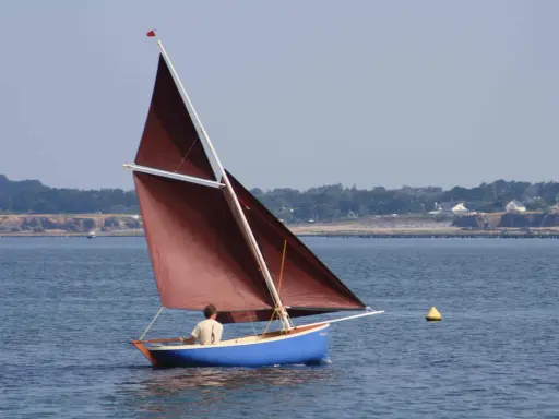 Un voilier bleu avec une voile marron navigue, tel une gazelle des îles, sur une mer calme. Au loin, une côte boisée bordée de maisons.