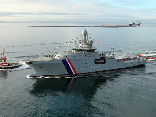 Un imposant bateau garde côte, le Landhelgisgæslan, navigue en mer, escorté par un remorqueur et suivi par des canots. Un hélicoptère survole la scène.