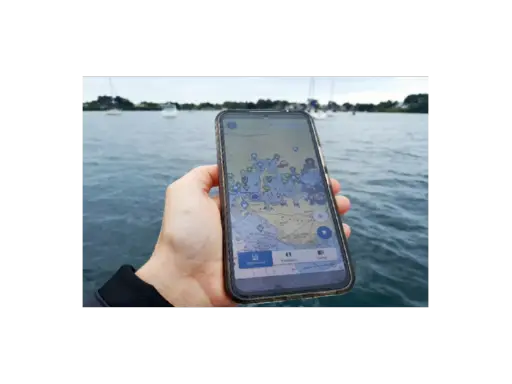 Main d'une personne tenant un smartphone affichant une carte marine avec des points d'intérêt. Le nav et co. sont visibles sur l'eau.