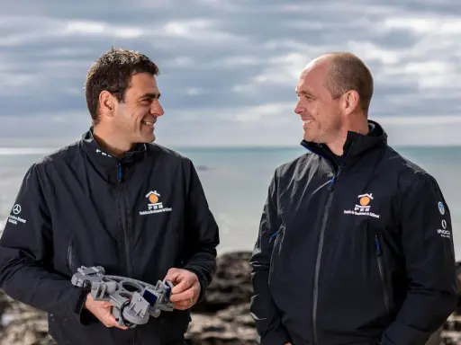 Damien Guillou et son collègue souriants sur la plage, l'un tenant un sextant, prêts pour une nouvelle aventure maritime.