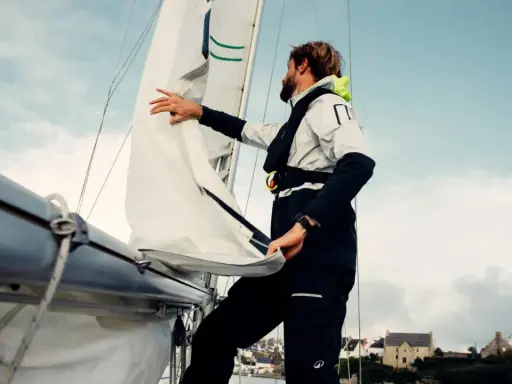 Un homme ajuste un type de voile sur un voilier, avec des maisons en arrière-plan.