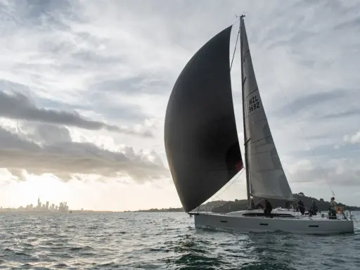 Un voilier navigue avec un grand gennaker noir gonflé par le vent, contrastant avec le spinnaker gris. La ville d'Auckland se dessine à l'horizon.