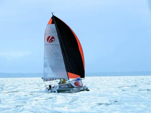 Voilier Heol 7.4 fendant les vagues, ses voiles bicolores gonflées par le vent. Deux marins à bord.