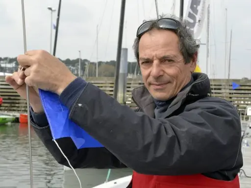 Marc Guillemot, skipper expérimenté, ajuste une voile bleue sur un ponton, prêt pour une nouvelle aventure en mer.