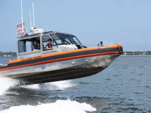 Un bateau du garde côte américain file sur l'eau, créant une gerbe d'écume. Deux membres d'équipage sont visibles à l'intérieur.