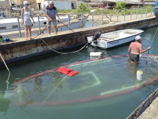 Un bateau accidenté est partiellement submergé dans un port. Des personnes sur le quai tentent de gérer la situation.