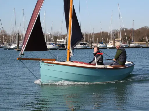 Deux hommes naviguent sur un petit bateau à voile bleu clair, avec d'autres voiliers en arrière-plan.