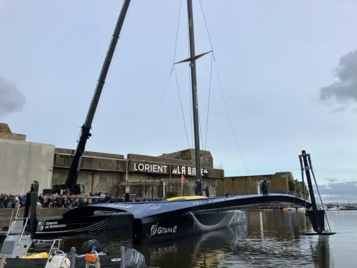Le trimaran Gitana 18, prêt pour l'aventure, est soulevé par une grue à Lorient. Une foule nombreuse est rassemblée pour admirer ce géant des mers.