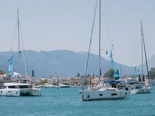 Trois voiliers naviguent dans une baie ensoleillée. Au premier plan, un catamaran monocoque blanc avec des voiles bleues et blanches. Derrière, un autre voilier blanc avec des personnes à bord. Au loin, une ville côtière et des montagnes.