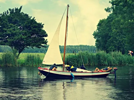 Un petit type de bateau à voile navigue sur un canal bordé d'arbres. Une personne se repose sur le pont, une autre est assise.