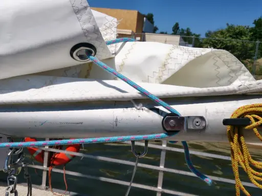 Le cordage bleu passe dans un œillet de la voile, prêt à ajuster la bosse de ris. Une amarre jaune est enroulée sur un taquet.