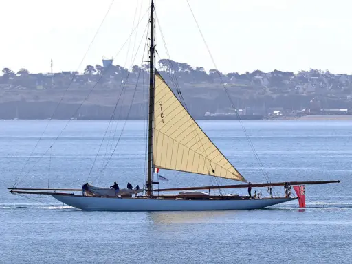 Un élégant voilier, surnommé "mariquita", navigue sur une mer bleue, sa grande voile beige déployée. Une côte verdoyante bordée de maisons se dessine à l'horizon.