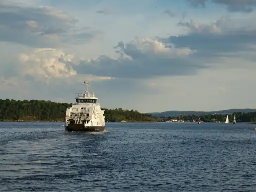Un ferry blanc navigue sur une eau bleue, laissant une traînée derrière lui. La vitesse du bateau est visible dans le sillage. Au loin, des voiliers et une côte boisée.