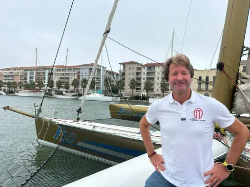 Un homme souriant sur un trimaran, peut-être le "Florence Arthaud", avec des bateaux et des immeubles en arrière-plan.