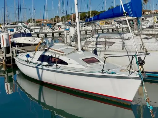 Un voilier blanc avec une voile bleue dans un port, reflété dans l'eau calme.
