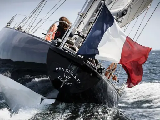 Le voilier Pen Duick VI fend les vagues, pavillon français au vent. Une silhouette se tient à la barre.