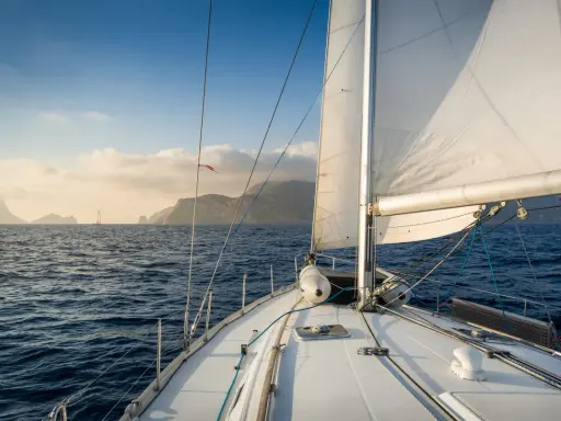 Le voilier navigue sur une mer bleue, le grand voile blanc gonflé par le vent. Au loin, des îles brumeuses se dessinent sous un ciel clair.