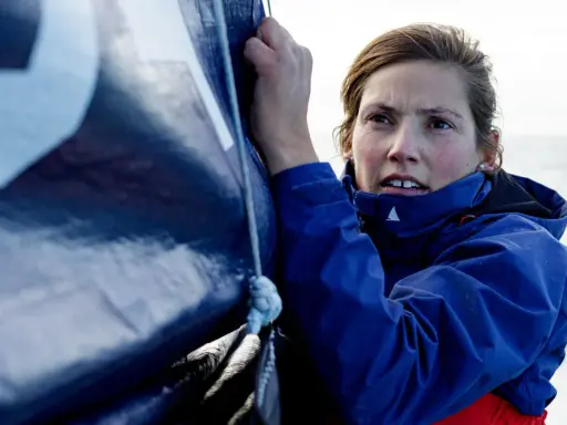 Tiphaine Ragueneau, concentrée, ajuste une voile bleue sur un bateau. La mer est visible en arrière-plan.