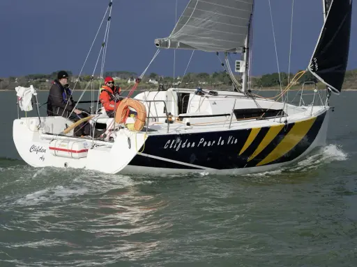 Voile rapide, le voilier "Clifden Pour la Vie" navigue sur une mer agitée. Deux personnes sont à bord, profitant du soleil.