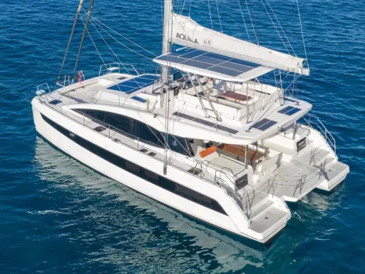 Un catamaran blanc, une véritable bicoque bateau, navigue sur une mer bleue. Ses voiles sont partiellement déployées, et des panneaux solaires brillent sur son toit.