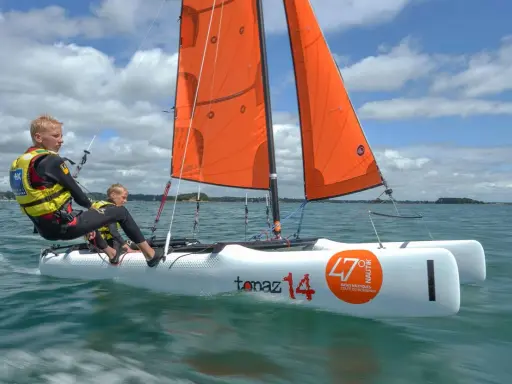 Deux jeunes garçons naviguent sur un petit voilier 2 places, leurs voiles oranges gonflées par le vent sous un ciel bleu et nuageux.