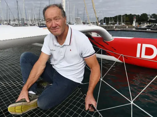 Francis Joyon, marin légendaire, pose sur le filet d'un trimaran rouge vif, prêt pour une nouvelle aventure en mer.