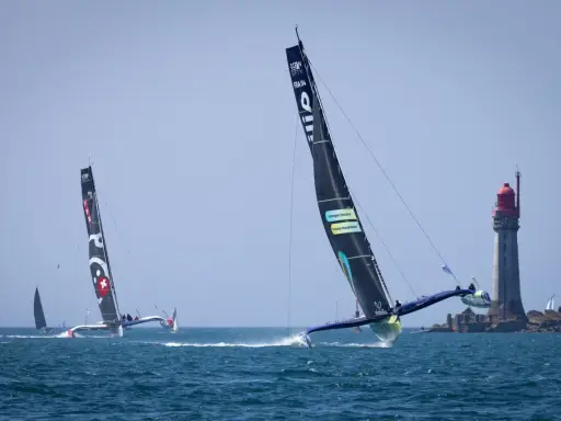Le trimaran Koesio fend les vagues, une course intense près d'un phare.