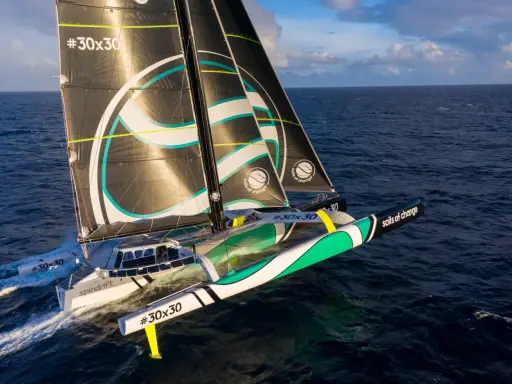 Un trimaran aux voiles noires et blanches, arborant le logo "sails of change", fend les vagues bleues sous un ciel nuageux.