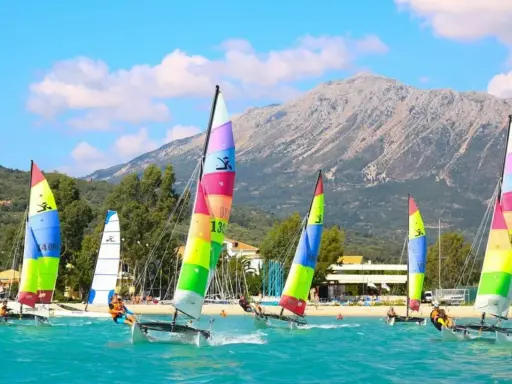 Plusieurs Hobie Cat 16 naviguent sur une mer turquoise, voiles multicolores, montagne en arrière-plan.