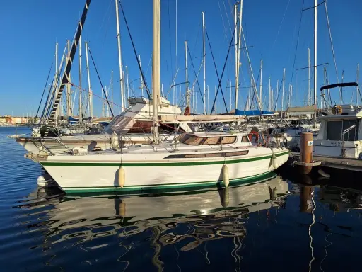 Un voilier blanc et vert, un Jeanneau Espace 1000, reflète sur l'eau calme d'un port.