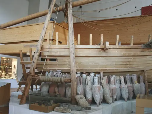 Amphores antiques alignées devant une reconstitution de navire en bois, rappelant le transport de vin par un ancien pinardier.
