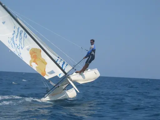 Un homme en trapeze sur un catamaran navigue sur une mer bleue sous un ciel clair. La voile arbore un dessin original.