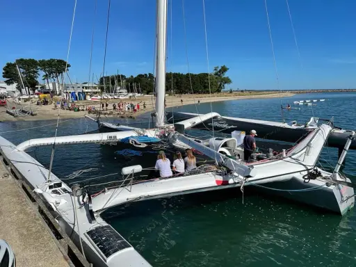 Le trimaran Flo, un bateau à trois coques, est amarré à quai. Des personnes sont à bord et sur la plage en arrière-plan.