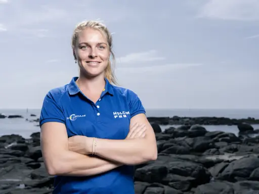 Rosalin Kuiper, souriante, pose les bras croisés devant un paysage côtier rocheux.