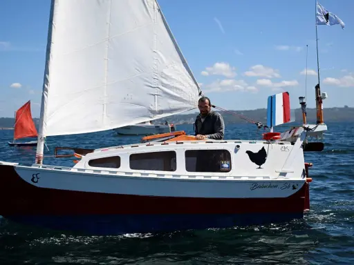 Un homme sourit sur son bateau nommé "Baluchon". Voiles blanches, coque rouge et bleue, drapeau français.