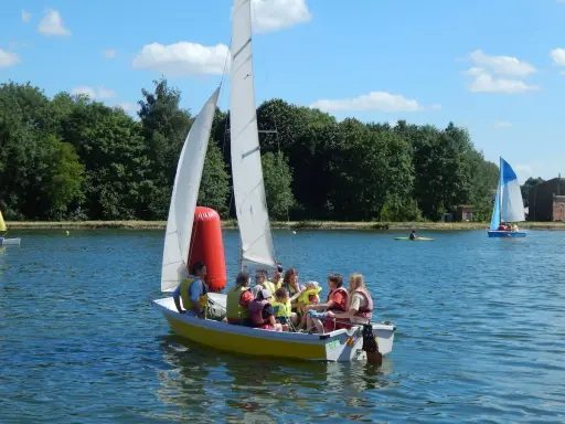 Un groupe de personnes, dont des enfants, profite d'une journée ensoleillée sur un bouchain bateau jaune. Un autre bateau à voile bleu navigue au loin.