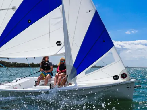 Deux jeunes femmes naviguent sur un voilier RS Venture par une journée ensoleillée. L'eau scintille sous le ciel bleu.