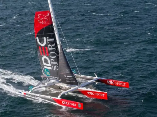 Le trimaran IDEC SPORT fend les vagues, une voile rouge et noire ornée d'un oiseau blanc.