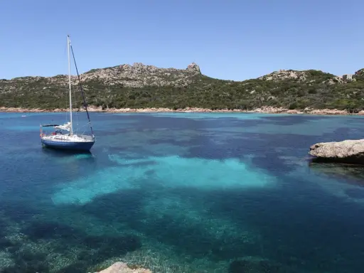 Un voilier bleu ancré dans les eaux turquoise de Cap Corse, avec des collines verdoyantes en arrière-plan sous un ciel clair.