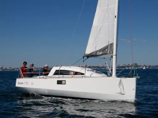 Un voilier Ikone 7.50 navigue par beau temps, avec trois personnes à bord. La voile est hissée, le ciel est bleu.