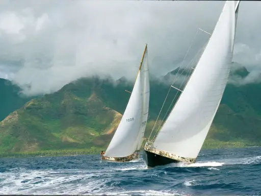 Deux voiliers de course, dont un avec le numéro 7020, naviguent sur une mer agitée, avec des montagnes verdoyantes et brumeuses en arrière-plan. Le pen duick 3 est en pleine action.