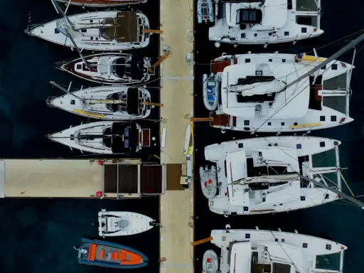 Vue aérienne d'un port avec plusieurs yachts à voile blancs amarrés, certains catamarans, et des annexes gonflables.