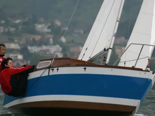 Couple en action sur un voilier transportable, naviguant sur des eaux agitées avec un village en arrière-plan.