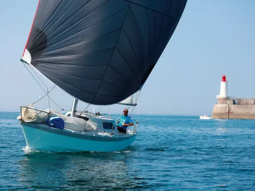 Un voilier bleu avec une grande voile noire navigue près d'un phare. Le skipper, portant un t-shirt bleu, semble observer un ovni 25 dans le ciel clair.