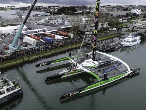 Le trimaran Sodebo Ultim 3, prêt pour la course, est amarré dans un port, entouré de spectateurs et de grues.
